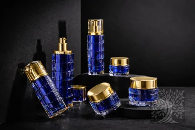 Cobalt blue acrylic skincare bottles and cosmetic jars with gold caps, staged on black stands with angled jar detail.